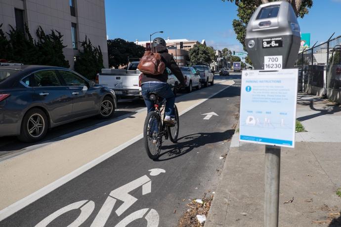 A person bikes on Valencia Street in a bike lane placed between a lane of parked cars and a sidewalk curb, with parking meters featuring instructional flyers.