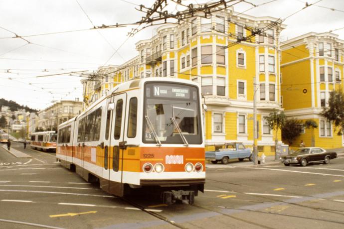 A Muni light rail vehicle with orange stripes and Muni worm logo crosses Church Street at Duboce in front of a yellow building.