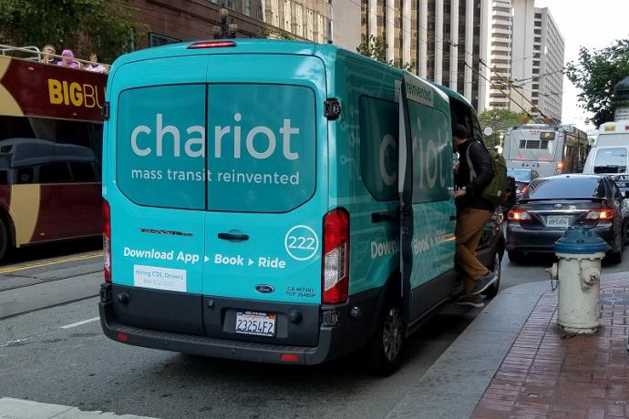 A shuttle van branded with the logo of Chariot, picks up a passenger near a fire hydrant on Market Street.