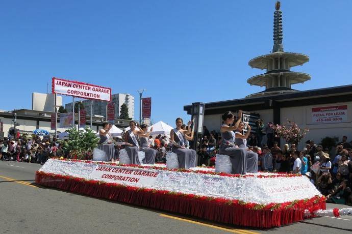 Ladies in gray dresses sit on a red and white float waving to the crowd under a bright blue sky.