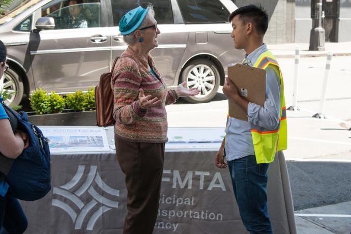 A woman talks with an SFMTA staffer holding a clipboard while standing on a sidewalk in front of a table that features the SFMTA logo.