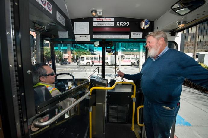 A Muni operator, seated in the bus driver seat, greets a customer with a smile.