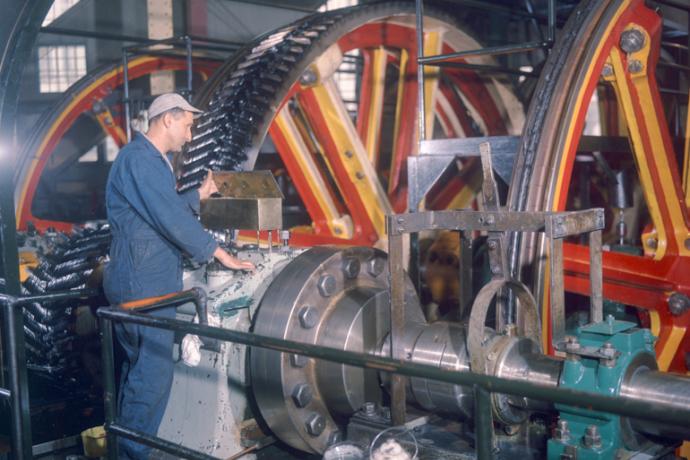 Color photo of a man standing next to the huge pulleys of the cable winding machinery, looking into a bronze oil box on top of a large bearing housing.