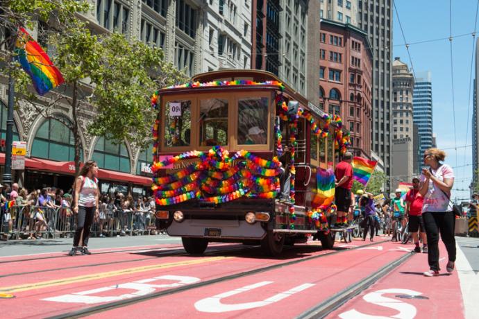 Color photograph from June 26, 2016 showing Muni motorized cable car 62 decorated in rainbow banners on Market Street as part of Pride parade.