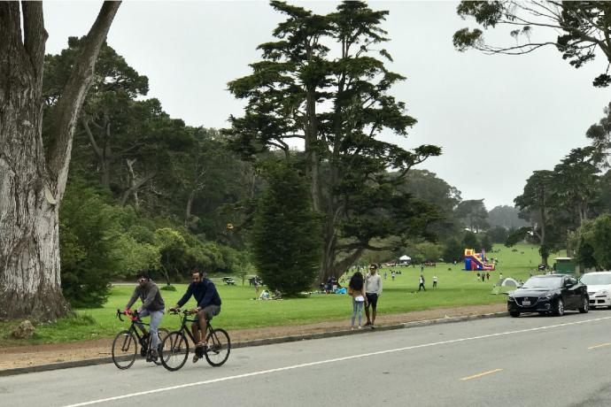 Image of people riding bicycles, a couple standing, and cars parked along John F. Kennedy Drive in Golden Gate Park.