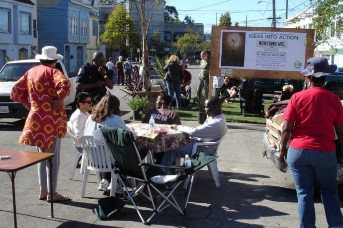 People gather at tables at a street meeting. Poster reads "Ideas into action".