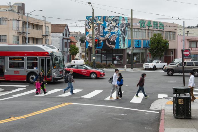Pedestrians, a taxi, and bus travel along Mission Street at Persia Ave