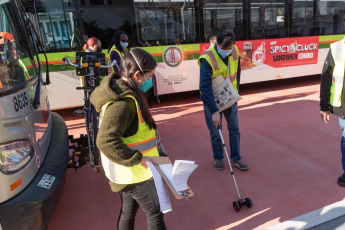 Two staff are shown taking measurements next to a Muni bus on the transit lane. One holds a rolling measuring device. The other 