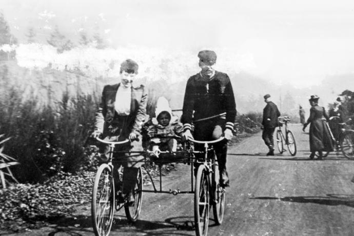 1890s photo of two people riding conjoined bicyles with a small child riding on a seat between the bicyles.