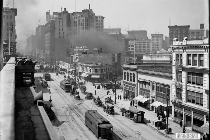 Black and white overhead view of street with horse-drawn and electric streetcars, horse drawn wagons, pedestrians, and buildings