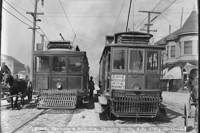 Two streetcars from 1911 shown with a person seen in the middle as well as a horse drawn carriage on the side.