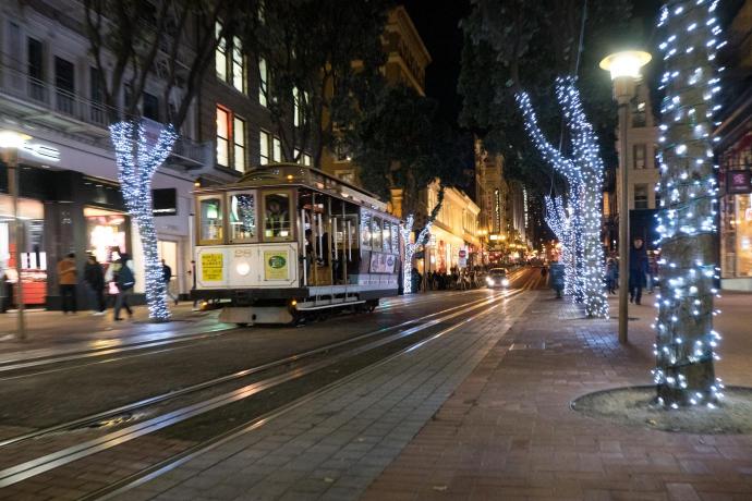 A Cable Car on a city street lined with buildings, shops and trees decorated with lights