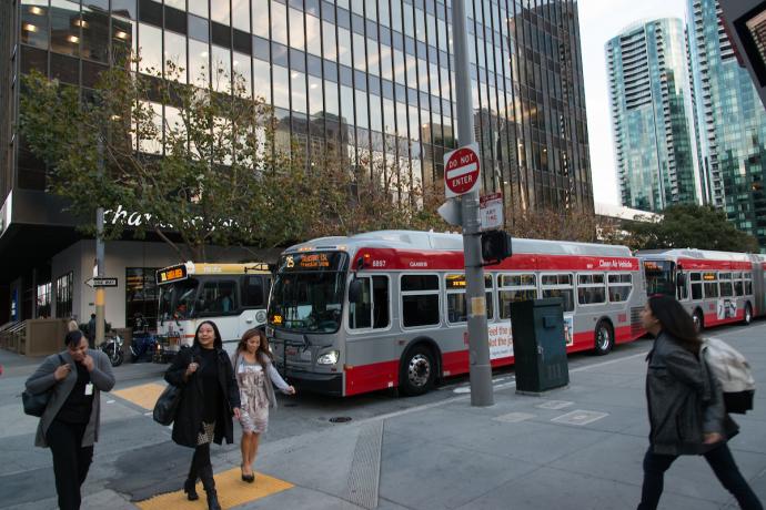 People crossing a downtown street in front of Muni and Golden Gate Transit buses