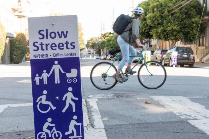 Close up image of a street sign with the words "Slow Streets" and multiple white figurine wheel users below. 