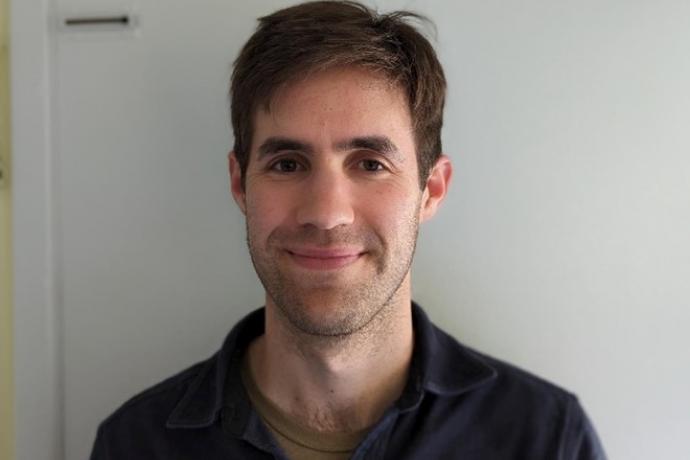 Man in a polo shirt smiling, standing in front of a white background wall.