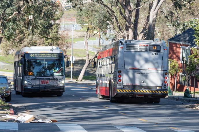 Two buses are side by side traveling in the opposite directions on a street lined with trees. 