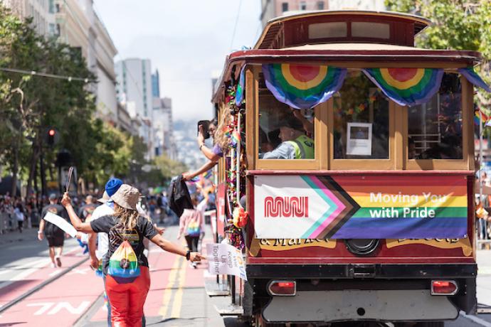 People march down the street at a parade around a historic cable car with rainbow flags.