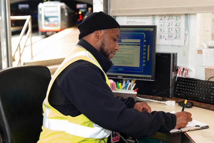 A photo of a Muni inspector writing on a clipboard at a desk with a computer and Muni Metro trains operating behind him.