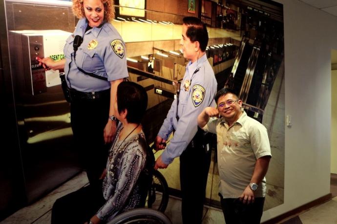 A man smiles in front of a picture of a three people with one of them in a wheelchair in front of an elevator.