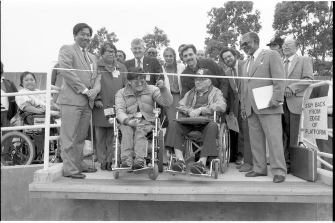 Black and white photo of people at a ribbon cutting ceremony. Two men in wheelchairs are in front to cut the ribbon.