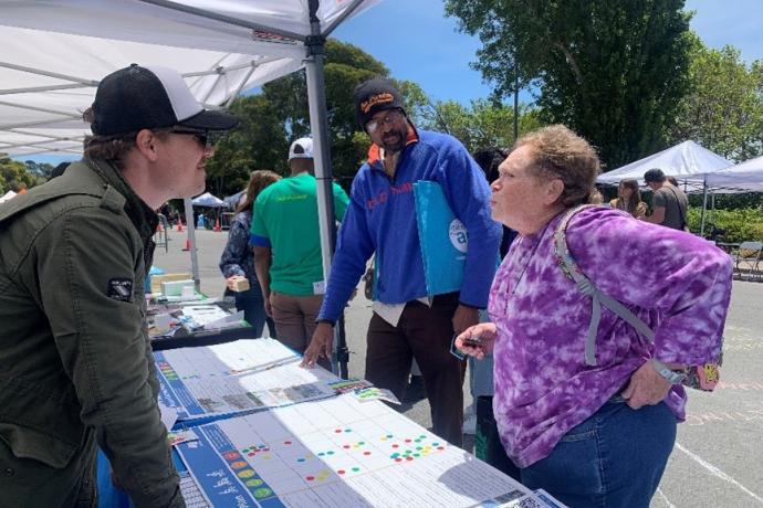 People talk underneath a tent with charts on a table and more people and tents in the background.