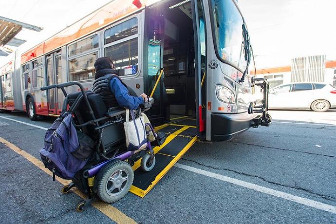 A person in a blue long sleeved shirt and black vest using a motorized wheelchair and using a ramp to board a Muni bus.