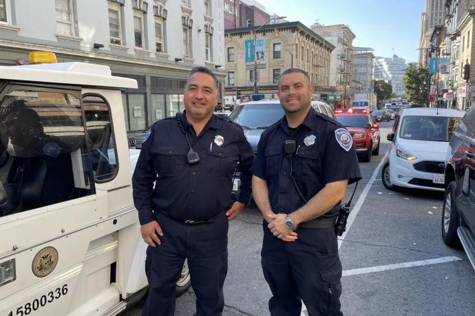 Two Parking Control Officers stand on the side of a busy street with cars and buildings in the background.