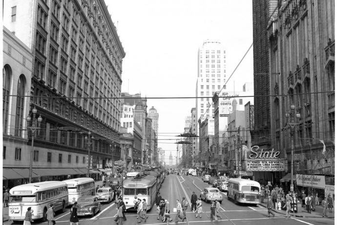 A black and white photo of a busy street with buses, cars and people crossing at a cross-walk.