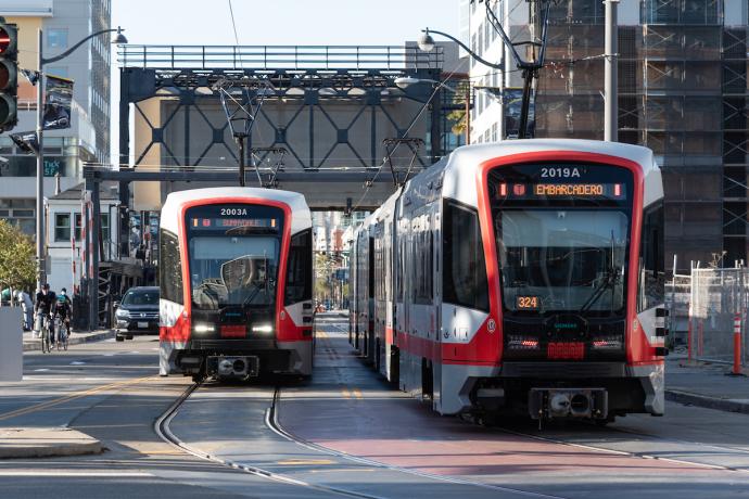 Two light rail trains pass each other on a street in San Francisco with a bridge hanging overhead in the background.
