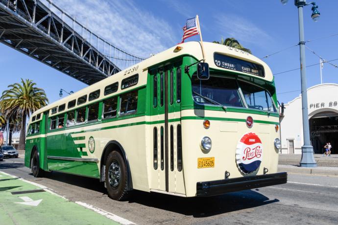 A vintage Muni bus that's green and cream in color passing under a bridge 