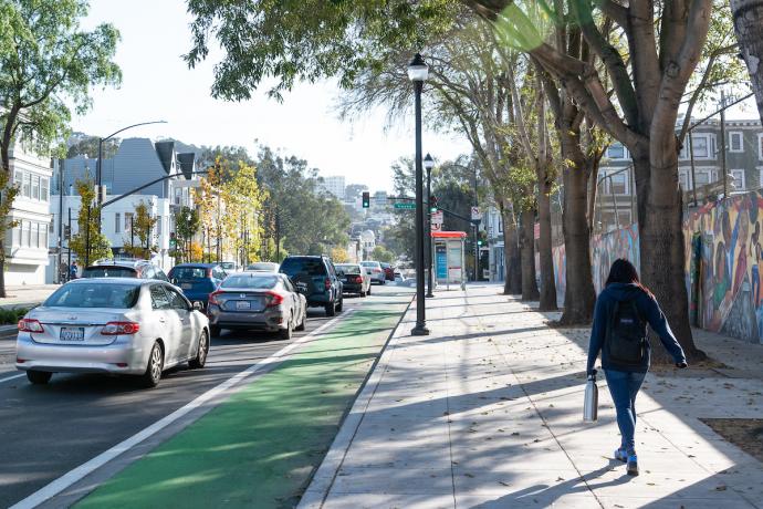 Woman walks on a sidewalk toward a bus shelter. On her left is a green bike lane beside lanes of cars.