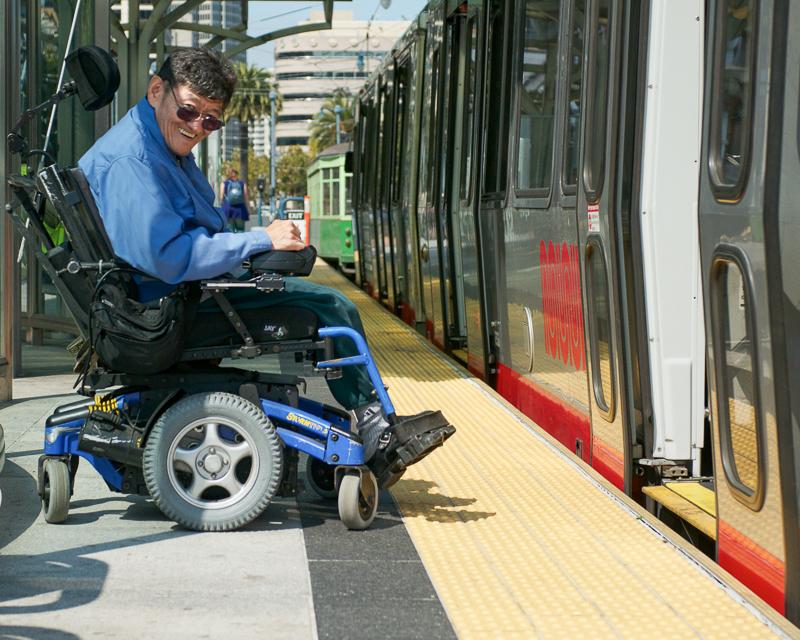 man in a wheelchair on boarding platform in front of open train doors, smiling and looking at the camera