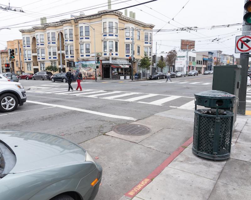 A car is parked behind a red zone at an intersection; pedestrians cross in front in a painted crosswalk