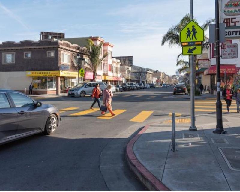 People cross in a painted crosswalk before a stopped car; to the right there's a bright neon sign with flashing lights below