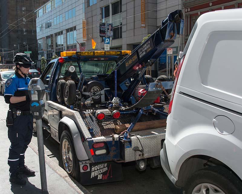 A parking control officer oversees the towing of a van from a parking-meter-regulated parking space