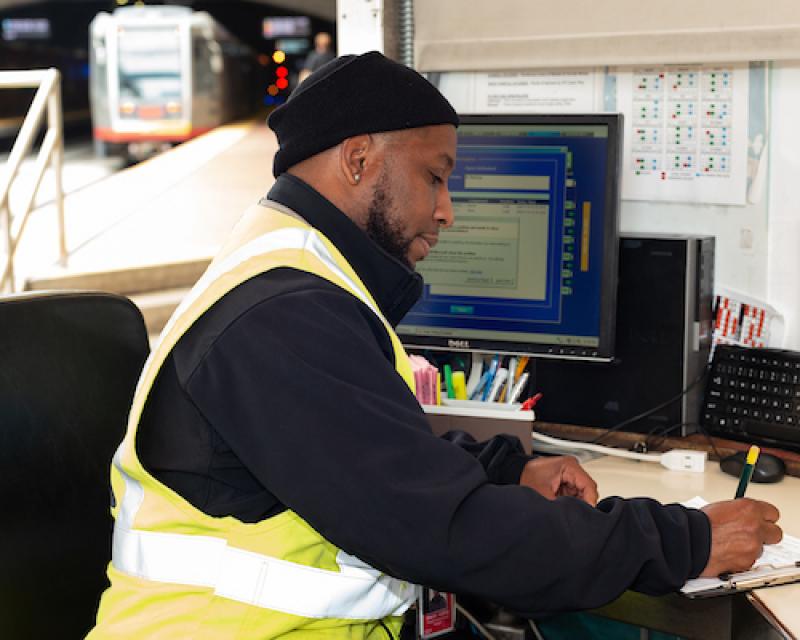 A photo of a Muni inspector writing on a clipboard at a desk with a computer and Muni Metro trains operating behind him.