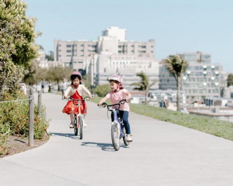 Two children riding bikes on a path with the San Francisco skyline in the background