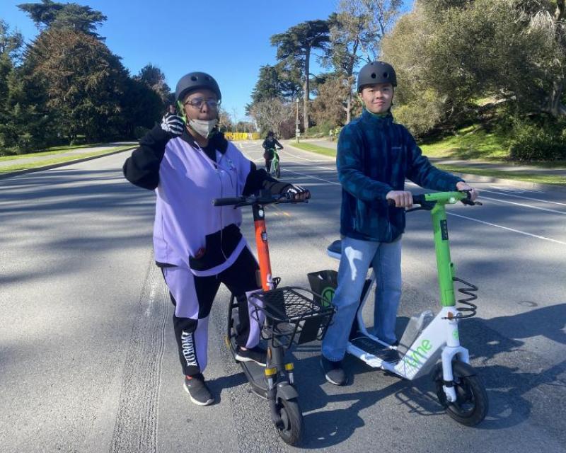 Two people with seated Spin and Lime scooters in Golden Gate Park; one is giving a thumbs up