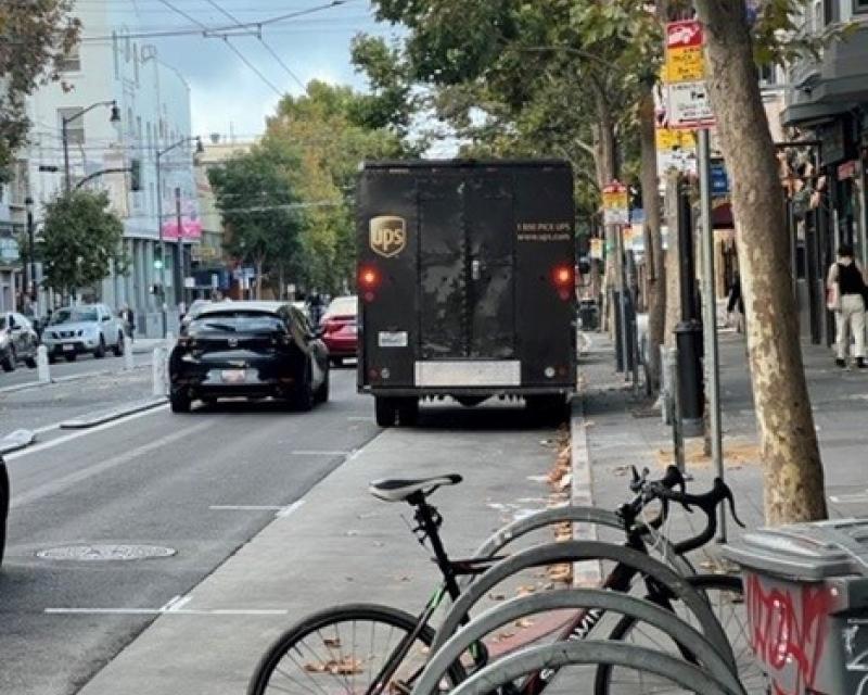 Parked bicycles on a sidewalk near cars and a delivery truck in a street. 