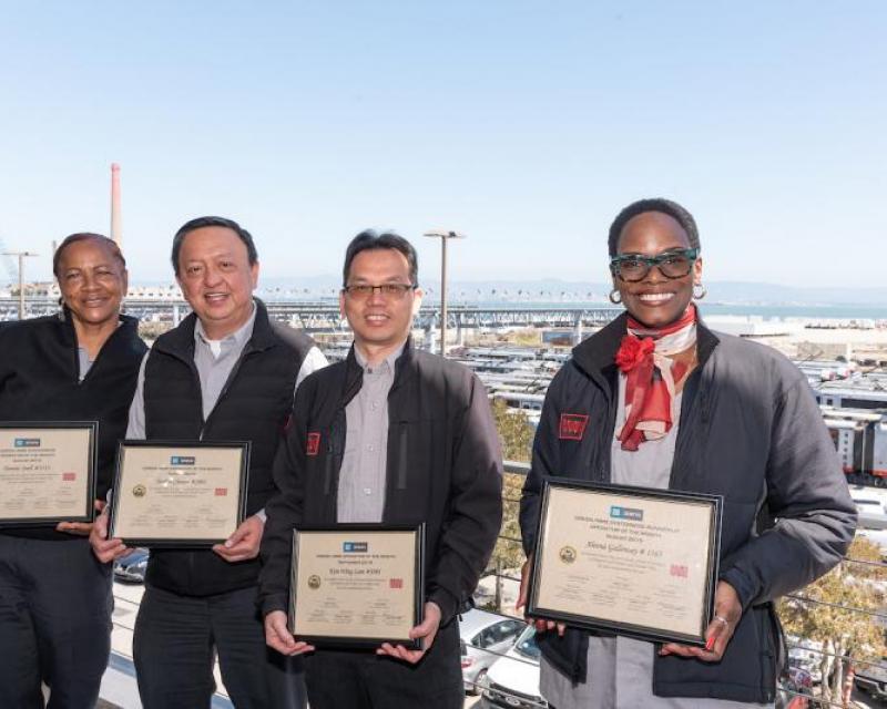 Four Muni operators stand, holding framed certificates with transit vehicles in the distant background.