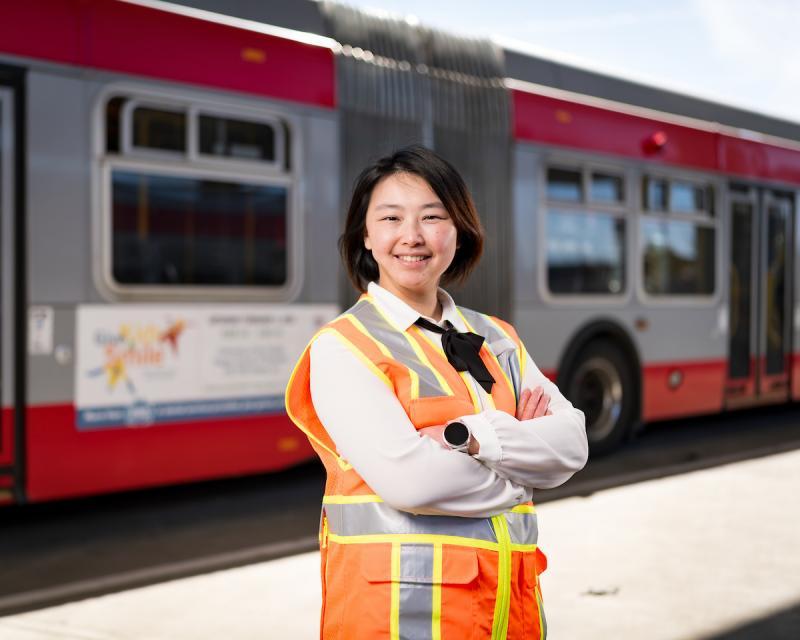 Becky Chen wears an orange safety vest, smiling and crossing her arms as she stands in front of a Muni bus.