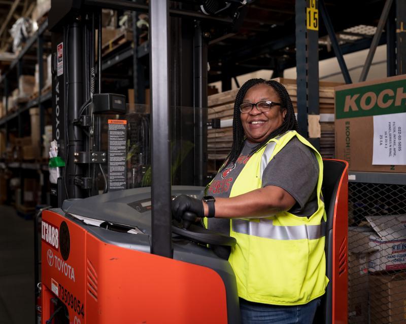 Evelyn smiles from her seat on a forklift. We see boxes of mechanical parts behind her.