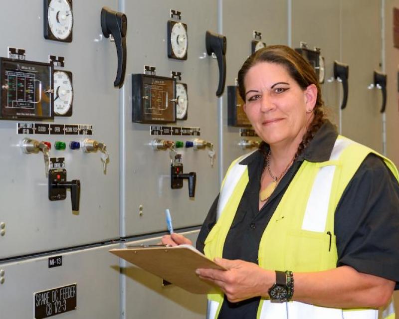 Ess records data from a substation. She wears a yellow vest and holds a clipboard as she smiles at the camera.