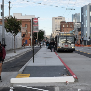 A man rides in a bike lane between the sidewalk and a concrete island where people board a Muni bus on 11th at Harrison.