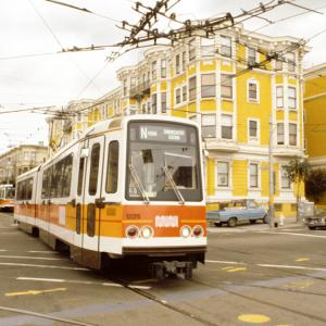 A Muni light rail vehicle with orange stripes and Muni worm logo crosses Church Street at Duboce in front of a yellow building.