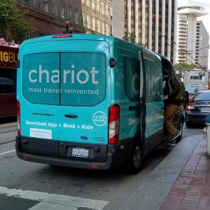 A shuttle van branded with the logo of Chariot, picks up a passenger near a fire hydrant on Market Street.