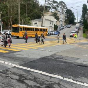 Students and crossing guards using the crosswalk, a bus is waiting to turn in the intersection 