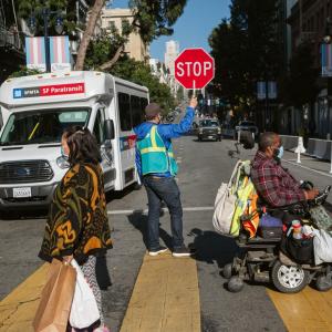 person in wheel chair and person walking, crossing street with crossing gaurd with Paratransit bus in background