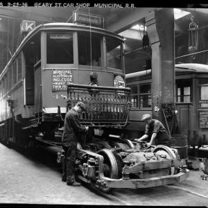 Inside the shops at Geary Car House, two mechanics work on a truck (the combined wheels, suspension, and motor of the streetcar)