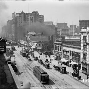 Black and white overhead view of street with horse-drawn and electric streetcars, horse drawn wagons, pedestrians, and buildings